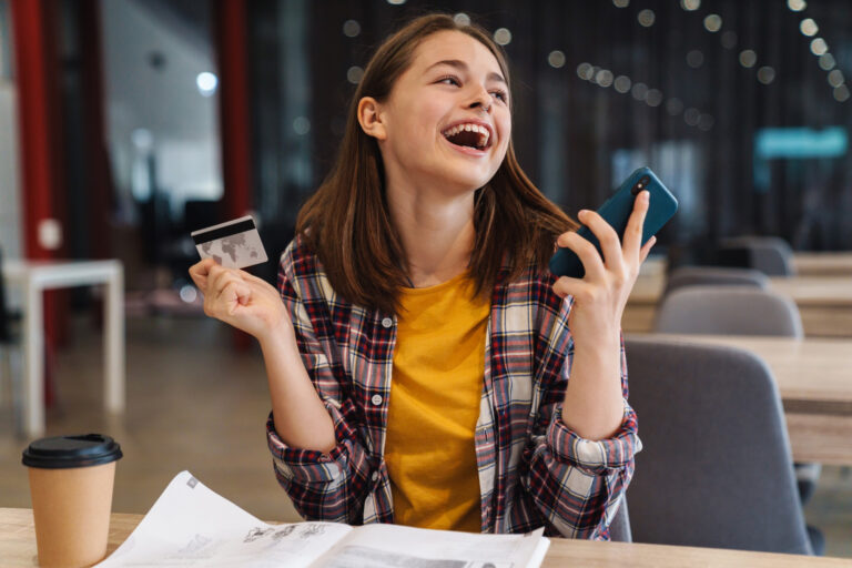 Portrait of delighted cute girl holding credit card and cellphone while doing homework in college library